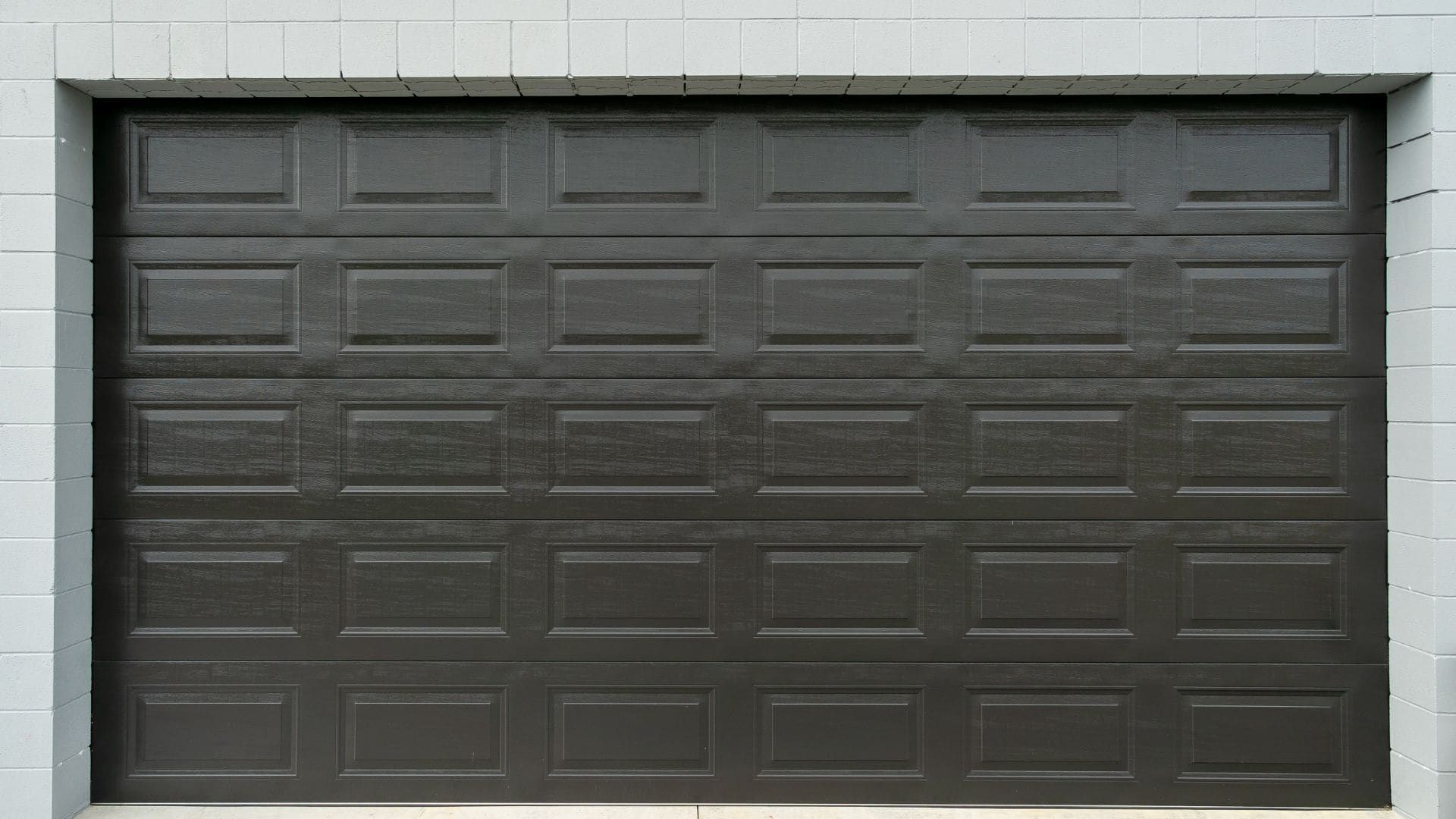 Black raised-panel double garage door on a grey block building
